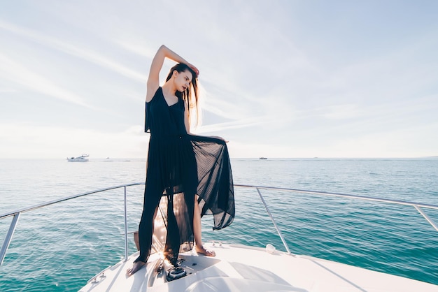 Uma linda garota luxuosa em um vestido preto de verão posando em seu iate branco desfrutando de uma viagem marítima