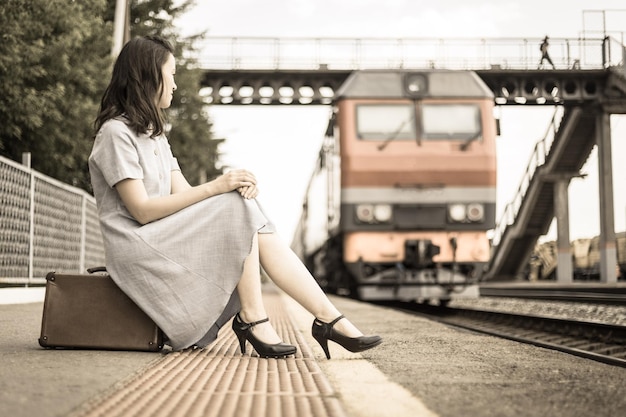 Foto uma linda garota na plataforma está sentada em uma mala no contexto de um trem que se aproxima processamento de estilo vintage o conceito de viajar de trem transporte ferroviário