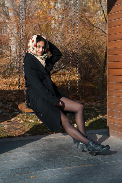 Uma linda garota sentada no parque em um balanço em sua cabeça é um lenço de cabeça tradicional russo