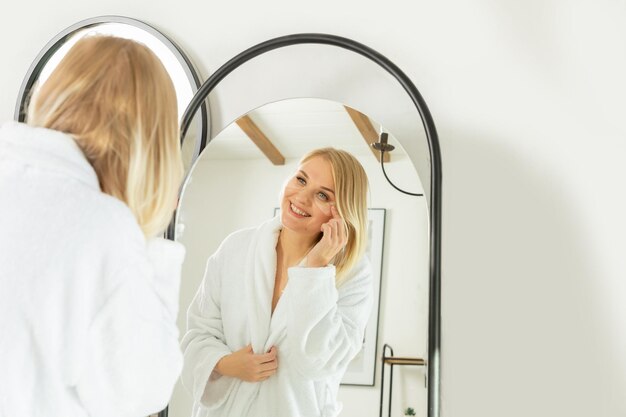 Foto uma linda garota sorridente em uma túnica branca está de pé na frente de um espelho cuidando de seu corpo