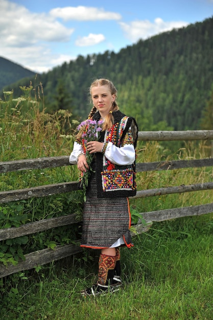 Uma linda garota ucraniana com flores nas mãos fica perto da cerca em um fundo de montanhas.