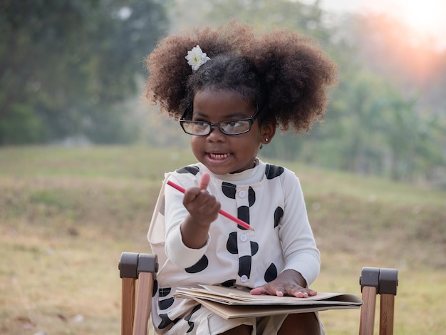 Uma linda garotinha afro-americana de óculos está sentada em uma cadeira e segurando um bloco de notas e um lápis em um parque público em uma bela manhãxa