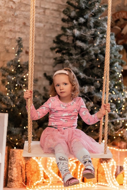 Uma linda garotinha com um elegante vestido rosa senta e rola em um balanço contra o fundo de árvores de natal e luzes em casa