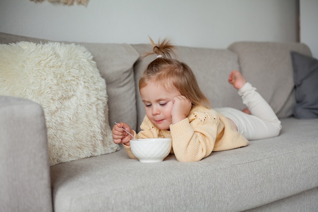 Uma linda garotinha de 2 anos com um suéter de tricô aconchegante está tomando café da manhã no sofá