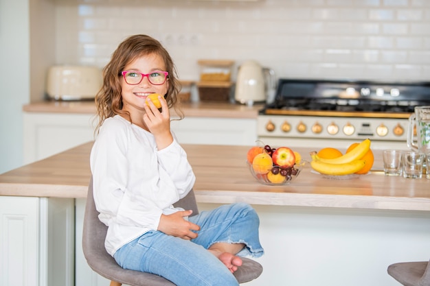 Uma linda garotinha de óculos está sentada na cozinha perto da mesa com frutas e segura um damasco nas mãos