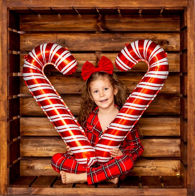 Uma linda garotinha de pijama de Natal segurando grandes doces de caramelo em um fundo de madeira na mão