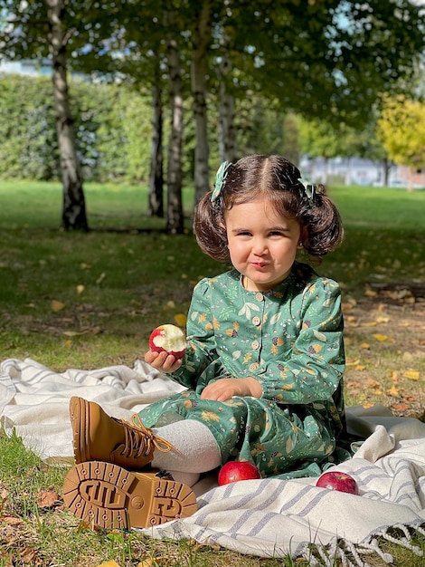 uma linda garotinha de vestido senta-se em uma manta de algodão no jardim de outono e come uma maçã vermelha