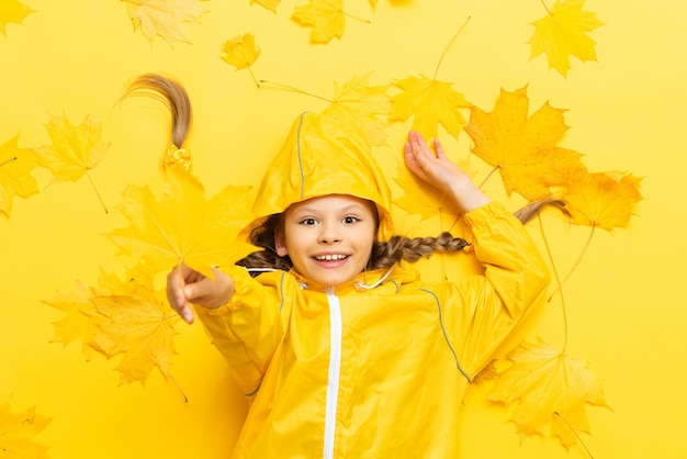 Uma linda garotinha em um fundo amarelo de outono com maple deixa uma criança em uma capa de chuva à prova d'água e um suéter de malha quente