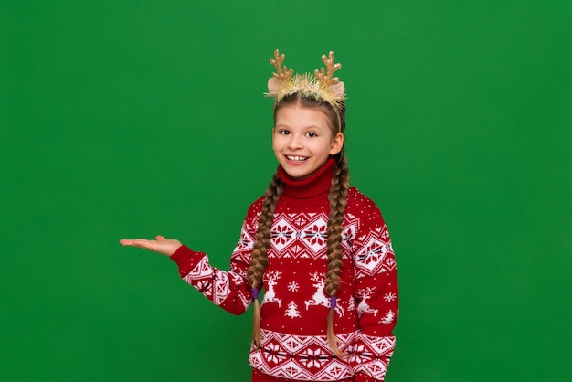 Uma linda garotinha em um suéter de Natal e cachecol com chifres de veado na cabeça tem um anúncio na mão um fundo verde isolado e um espaço de cópia