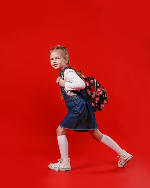 Uma linda garotinha em um vestido jeans com uma mochila nos ombros caminha sobre um fundo vermelho isolado.