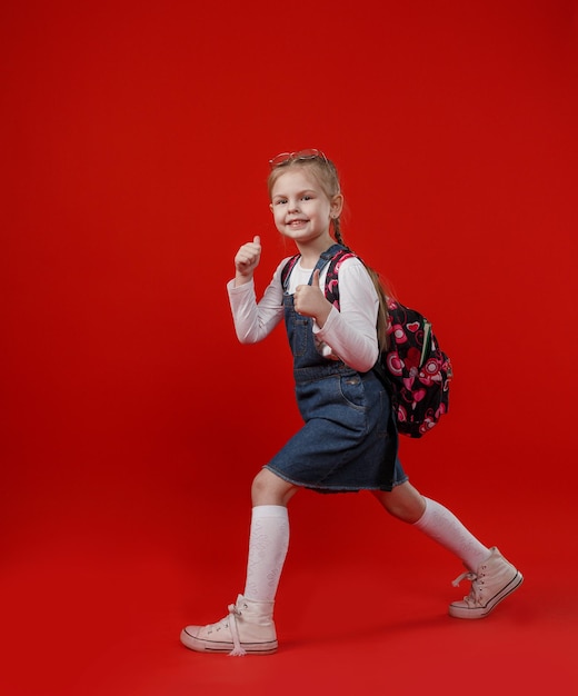 Uma linda garotinha em um vestido jeans com uma mochila nos ombros caminha sobre um fundo vermelho isolado.