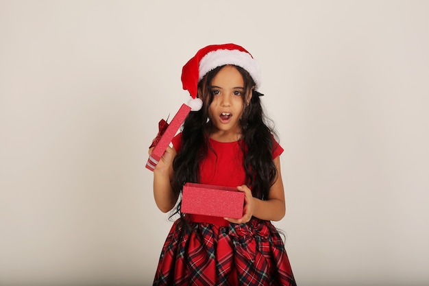 uma linda garotinha em um vestido vermelho e um chapéu de Papai Noel segurando uma caixa de presente nas mãos