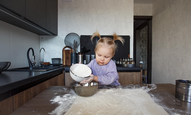 Uma linda garotinha está aprendendo a cozinhar na cozinha. A criança derrama farinha na mesa da cozinha