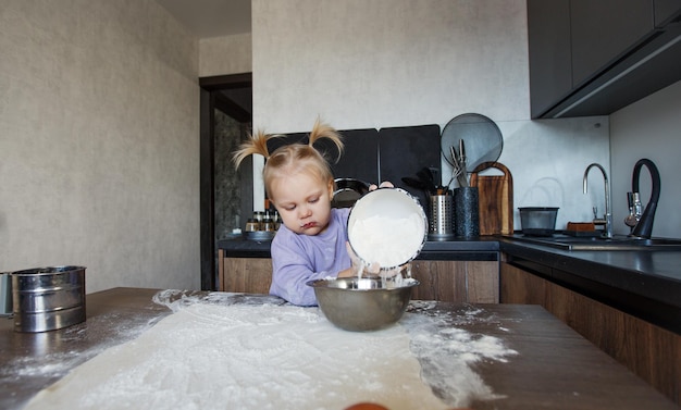 Uma linda garotinha está aprendendo a cozinhar na cozinha. A criança derrama farinha na mesa da cozinha