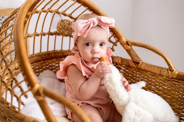 Uma linda garotinha europeia com olhos castanhos em uma roupa rosa em um berço de vime infantil no quarto das crianças em casa