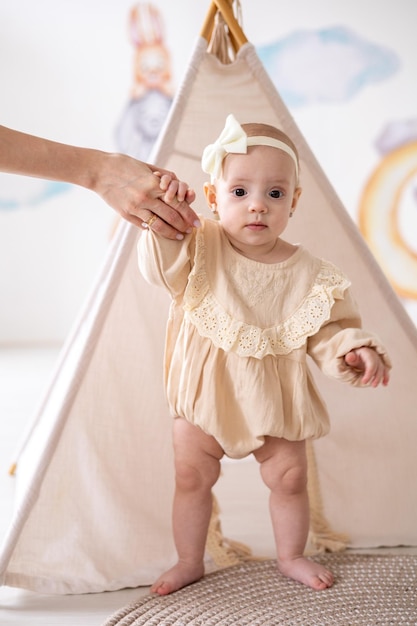 Uma linda garotinha europeia de olhos castanhos em um macacão bege feito de tecido natural dá seus primeiros passos pela mão com a mãe no quarto das crianças em casa