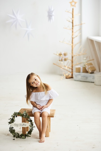 Uma linda garotinha sentada em um banquinho de madeira em um lindo vestido no quarto branco