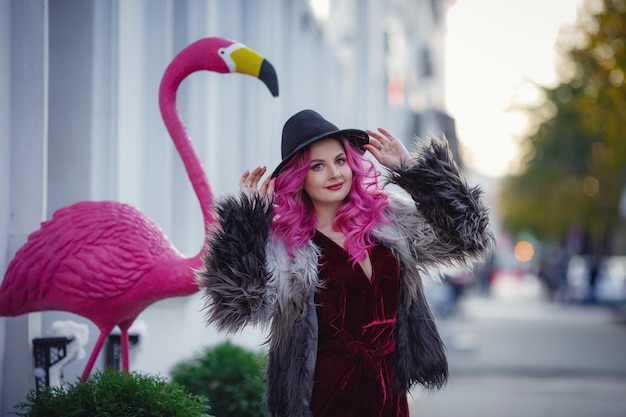 Uma linda jovem com cabelo rosa e um casaco incomum anda pela cidade à noite desfruta da luz das guirlandas e da luz do sol