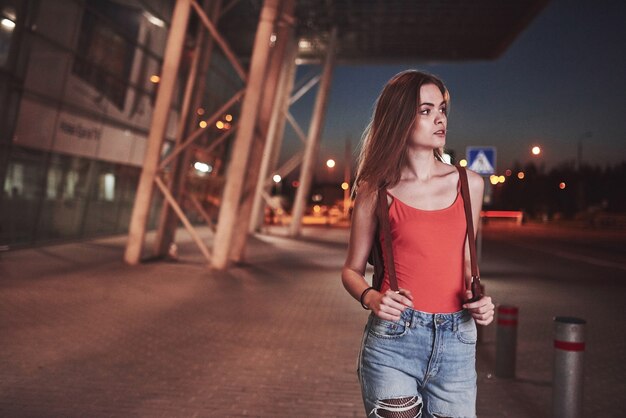 Uma linda jovem com uma mochila atrás dos ombros está de pé na rua perto de um aeroporto ou estação ferroviária em uma noite quente de verão. ela acabou de chegar e espera um táxi ou seus amigos.