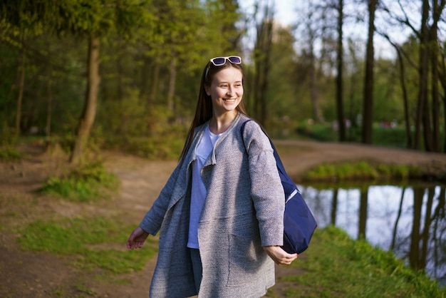 Uma linda jovem de óculos escuros e carregando uma bolsa caminha em um parque florestal perto do lago em um dia ensolarado