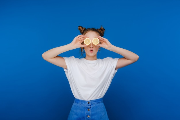 Uma linda jovem de pé sobre um fundo azul tem limões nas mãos e cobre os olhos com eles.