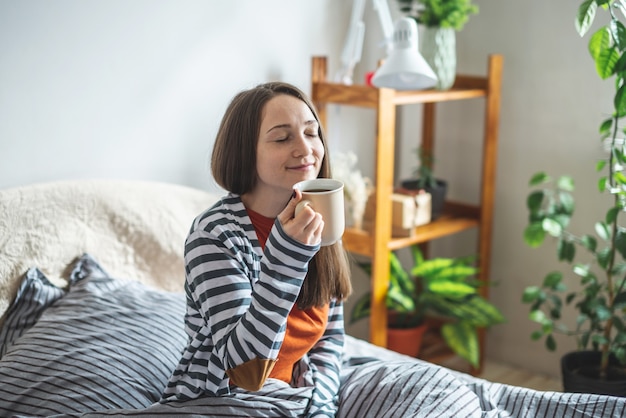 Uma linda jovem de pijama está sentada na cama em uma manhã ensolarada tomando uma xícara de café