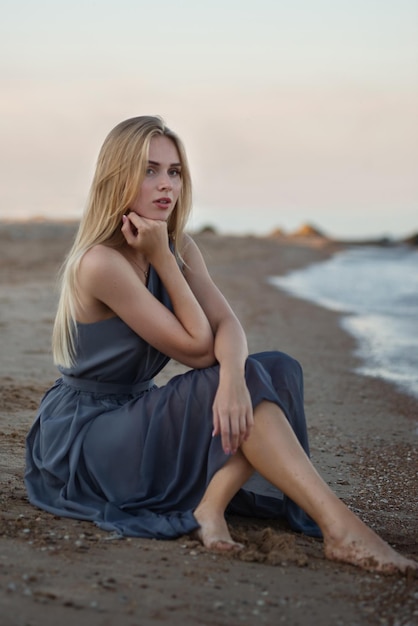 Uma linda jovem em um longo vestido azul senta-se à beira-mar e observa o pôr do sol