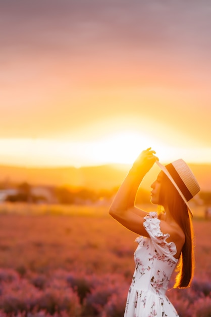 Uma linda jovem em um vestido delicado e chapéu caminha por um belo campo de lavanda e aprecia o aroma das flores. Férias e beleza natural.