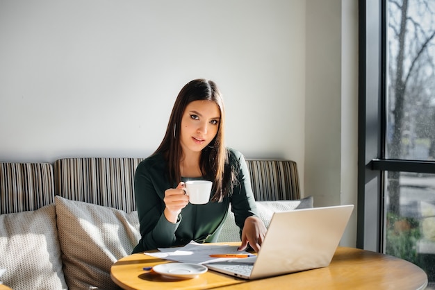 Uma linda jovem está sentada em um café, trabalhando em um computador e se comunicando em redes sociais.