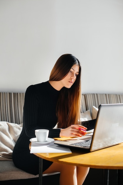 Uma linda jovem está sentada em um café, trabalhando em um computador e se comunicando em redes sociais.