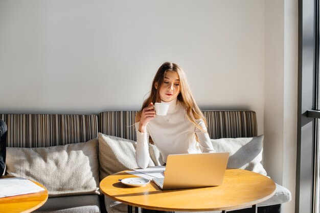 Uma linda jovem está sentada em um café, trabalhando em um computador e se comunicando em redes sociais.