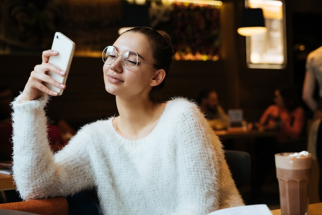 Uma linda jovem estudante de óculos sentada no café após o estudo, olhando para o smartphone