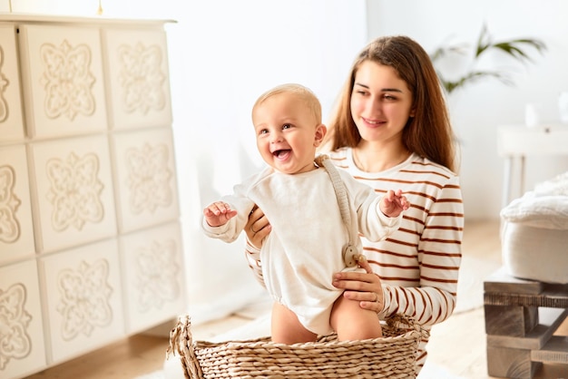 Uma linda jovem mãe está segurando um bebê de pé em uma cesta e rindo