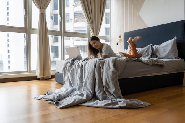 Uma linda jovem morena está trabalhando no laptop enquanto estava deitado em uma cama por uma janela panorâmica. interior moderno e elegante. um local de trabalho acolhedor