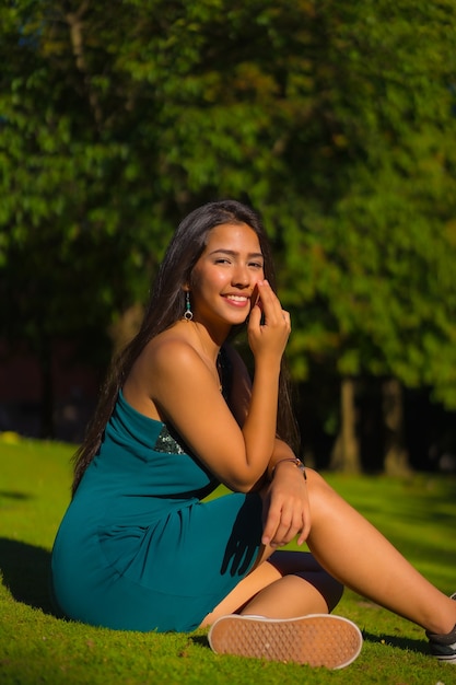 Uma linda jovem morena latina com cabelos longos e lisos, usando um vestido verde justo. Retrato de uma pessoa sentada na grama de um parque no verão