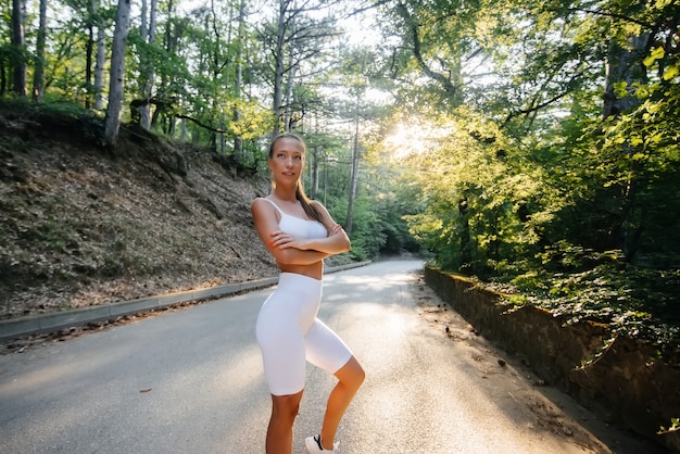 Uma linda jovem posa antes de correr o treinamento, na estrada em uma floresta densa, durante o pôr do sol. Um estilo de vida saudável e correr ao ar livre.