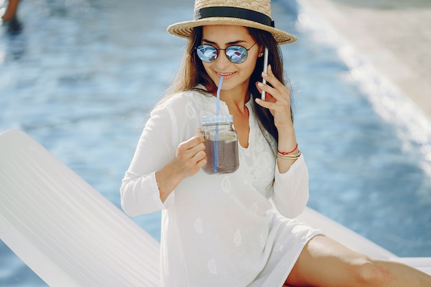 Uma linda jovem sentado em uma esplanada à beira da piscina com telefone e cocktail