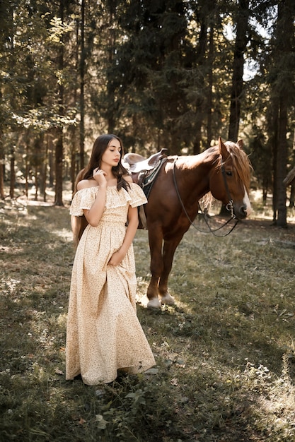 Uma linda jovem vestida com um vestido fica perto de um cavalo na floresta