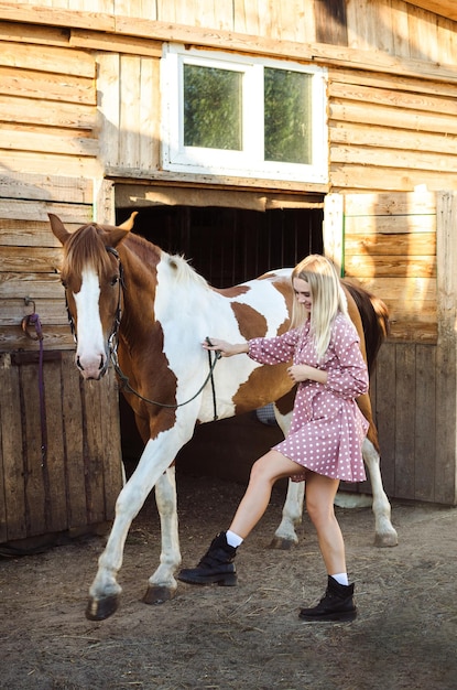 Uma linda loira está brincando com seu cavalo vermelho e branco no estábulo em pleno crescimento