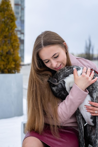 uma linda loira está sentada em um banco na cidade e abraçando um cachorro whipet