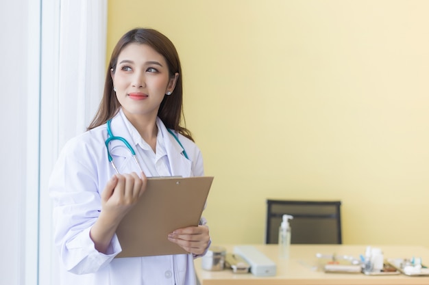 Uma linda médica asiática sorrindo, segurando uma prancheta e olhando pela janela.