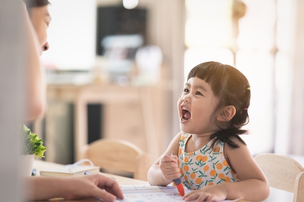 Uma linda menina asiática desenhando com papel branco na mesa de madeira em casa com a mãe