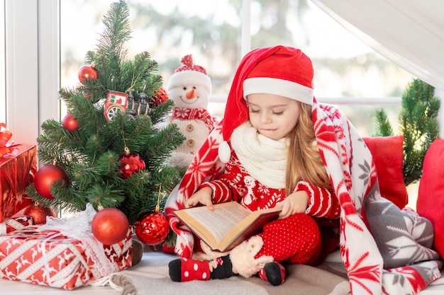 Uma linda menina está lendo um livro sentado no parapeito da janela na janela da casa na árvore de Natal e esperando o ano novo ou o Natal com um chapéu de Papai Noel vermelho e sorrindo