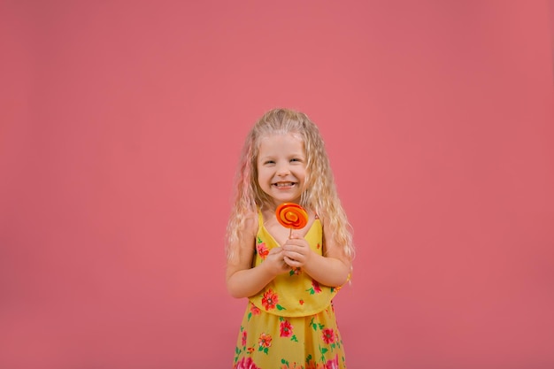 Foto uma linda menina loira em um vestido amarelo com um pirulito em um fundo rosa