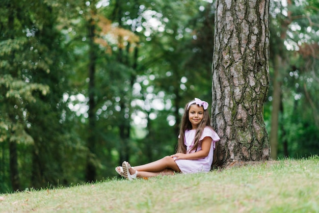 Uma linda menina sentada perto de uma árvore relaxando desfrutando de um dia ensolarado em um parque verde Férias escolares de verão