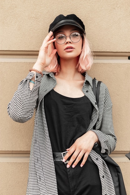 Uma linda modelo de mulher adolescente com óculos em um boné estiloso com uma camisa xadrez casual em uma camiseta preta fica perto de uma parede bege na rua e olha para a câmera