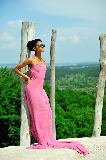 Uma linda modelo nigeriana de vestido rosa fica em óculos de sol em uma montanha e olha para o sol