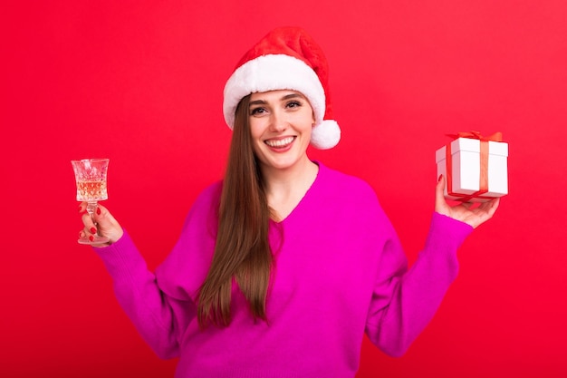 Uma linda morena com um chapéu de Papai Noel bebe champanhe e segura uma caixa de presente em um fundo vermelho
