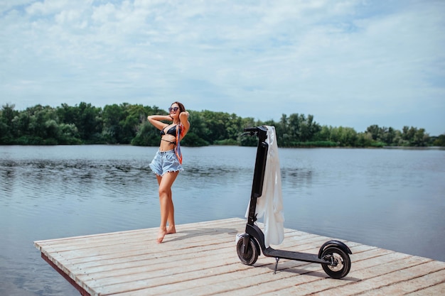 Foto uma linda morena de maiô toma banho de sol em uma ponte de madeira e há uma scooter por perto. conceito de ecologia e saúde. foto de alta qualidade