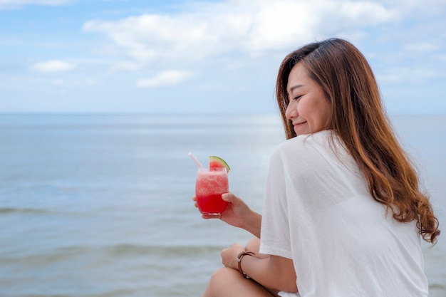 Uma linda mulher asiática bebendo suco de melancia enquanto está sentada à beira-mar